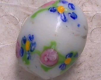 May include: An oval-shaped white glass bead featuring a floral pattern with blue petals, yellow centers, pink circular accents, and green stems. The surface of the bead is smooth and glossy, displaying a multi-colored design embedded within the glass.