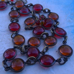 Puede incluir: Un collar vintage con cabujones redondos de vidrio ámbar y rojo. Las piedras de vidrio están engarzadas en una cadena de metal color bronce. El collar se exhibe sobre una superficie azul claro.
