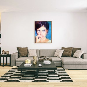 May include: A framed portrait of a woman with dark hair and blue eyes. The painting has a soft, impressionistic style with a blue and beige colour palette. The artwork hangs on a white wall above a grey sectional sofa in a modern living room.