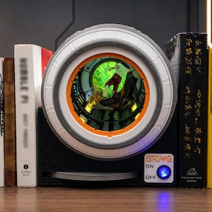 May include: A black bookend featuring a circular, light-up porthole display with a green, glowing interior scene. The base includes a white label with the text ASTROPHAGE, an ON/OFF switch with a blue light, and a series of thin, stacked circular discs.
