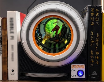 May include: A black bookend featuring a circular, light-up porthole display with a green, glowing interior scene. The base includes a white label with the text ASTROPHAGE, an ON/OFF switch with a blue light, and a series of thin, stacked circular discs.