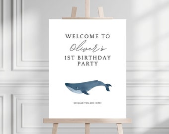 May include: A white welcome sign with blue whale illustration. The sign reads "Welcome to Oliver's 1st Birthday Party. So glad you are here!"