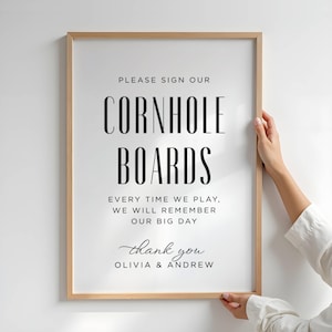 May include: A vertical rectangular sign in a light wood frame with black text on a white background that reads: PLEASE SIGN OUR CORNHOLE BOARDS EVERY TIME WE PLAY, WE WILL REMEMBER OUR BIG DAY thank you OLIVIA & ANDREW in a mix of serif and script fonts.