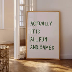 May include: A framed print with the text "ACTUALLY IT IS ALL FUN AND GAMES" in green letters on a white background. The frame is a light brown color, and the print is leaning against a wall.