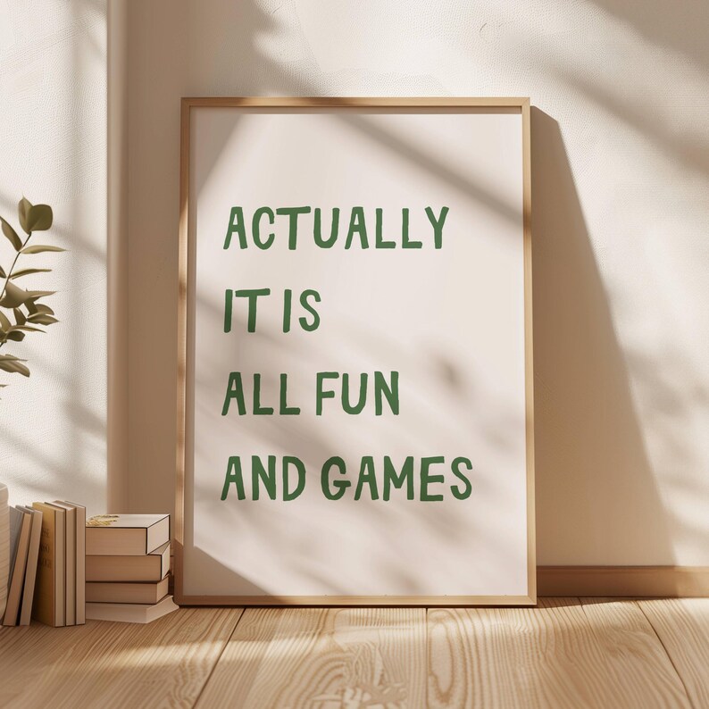 May include: A framed print with the text "ACTUALLY IT IS ALL FUN AND GAMES" in green letters. The print is leaning against a wall, with a stack of books and a plant visible in the foreground. The frame is a light brown color.