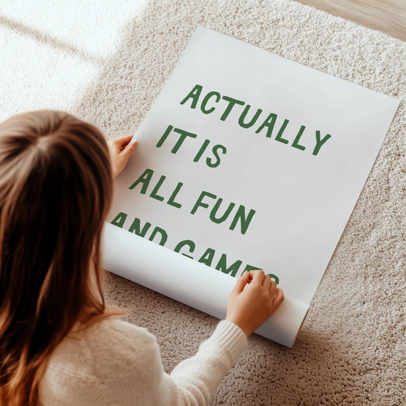 May include: A white poster with the text "ACTUALLY IT IS ALL FUN AND GAMES" in green lettering. The poster is being unrolled on a beige carpet. The image is taken from an overhead angle.
