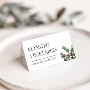 May include: A white folded card with the words "ROASTED VEGETABLES" in black, with "VEGETARIAN / GLUTEN FREE" below. The card is decorated with a watercolor holly and berry design. The card is on a white plate.