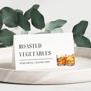 May include: White card with black text that reads "ROASTED VEGETABLES" and "VEGETARIAN / GLUTEN FREE". Two glasses of amber liquid with ice cubes are pictured on the right side of the card.