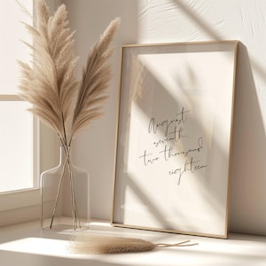 May include: A gold framed print with the text "August seventh, two thousand eighteen" on a white background. The print is sitting on a white surface with a vase of dried pampas grass.
