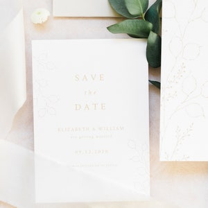 Wedding stationery flat lay with close up of minimal save the date card. Design is printed in pale gold on luxury white textured paper. Design features delicate hand drawn winter botanicals, honesty and seeds.