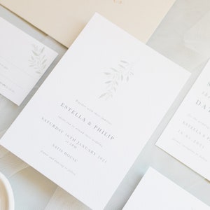 Wedding stationery suite flat lay with 5x7 invitation, A6 save the date, details card, A7 rsvp card, neutral envelope with calligraphy addressing. Minimal watercolour branch in pale green. Modern serif font for all text in grey. Printed on luxe paper