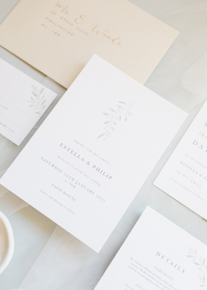 Wedding stationery suite flat lay with 5x7 invitation, A6 save the date, details card, A7 rsvp card, neutral envelope with calligraphy addressing. Minimal watercolour branch in pale green. Modern serif font for all text in grey. Printed on luxe paper