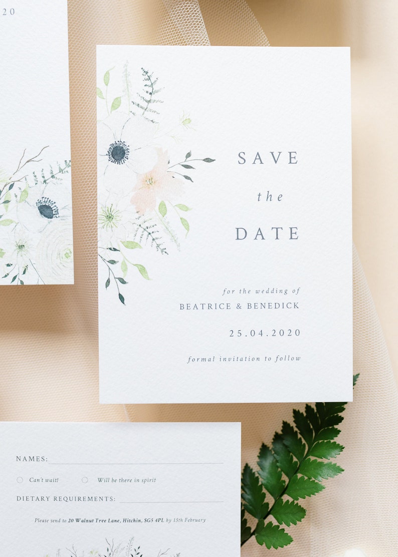 Wedding stationery flat lay with A6 portrait save the date card. Design features spring watercolour florals in peach, white and cream with anemone and ranunculus. Minimal design with elegant serif font printed on luxury white textured card stock.