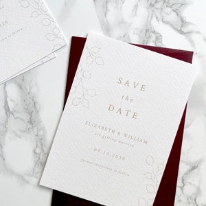 Wedding save the date card with burgundy envelope. Modern and minimal design with hand drawn honesty branches in opposite top and bottom corner. Text is centred. Printed in pale gold on luxury white textured paper.