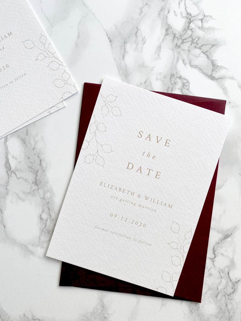 Wedding save the date card with burgundy envelope. Modern and minimal design with hand drawn honesty branches in opposite top and bottom corner. Text is centred. Printed in pale gold on luxury white textured paper.