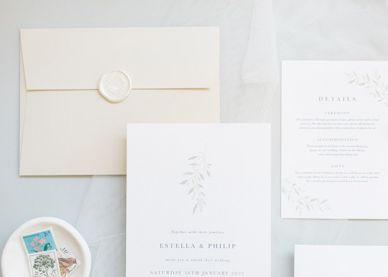 Wedding stationery flat lay with 5x7 invitation, A6 details card, stone envelope and white wax seal. Minimal design with watercolour eucalyptus branch at top with minimal text below in grey, classic serif font. Printed on luxury white textured paper