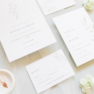 Wedding stationery suite flat lay with 5x7 portrait invitation, A6 portrait save the date, details card, A7 rsvp card. Minimal watercolour foliage design in pale green. Modern serif font for all text in grey. Printed on luxury white textured paper.