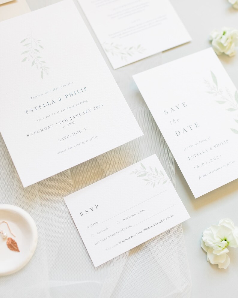 Wedding stationery suite flat lay with 5x7 portrait invitation, A6 portrait save the date, details card, A7 rsvp card. Minimal watercolour foliage design in pale green. Modern serif font for all text in grey. Printed on luxury white textured paper.