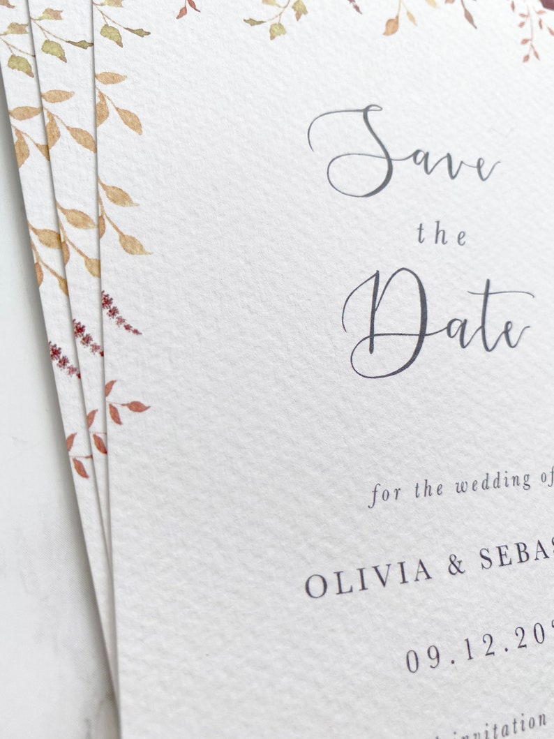 Close up of save the date pile, taken at an angle to see texture of the luxury paper. Featuring watercolour autumn leaves and modern calligraphy printed in grey.