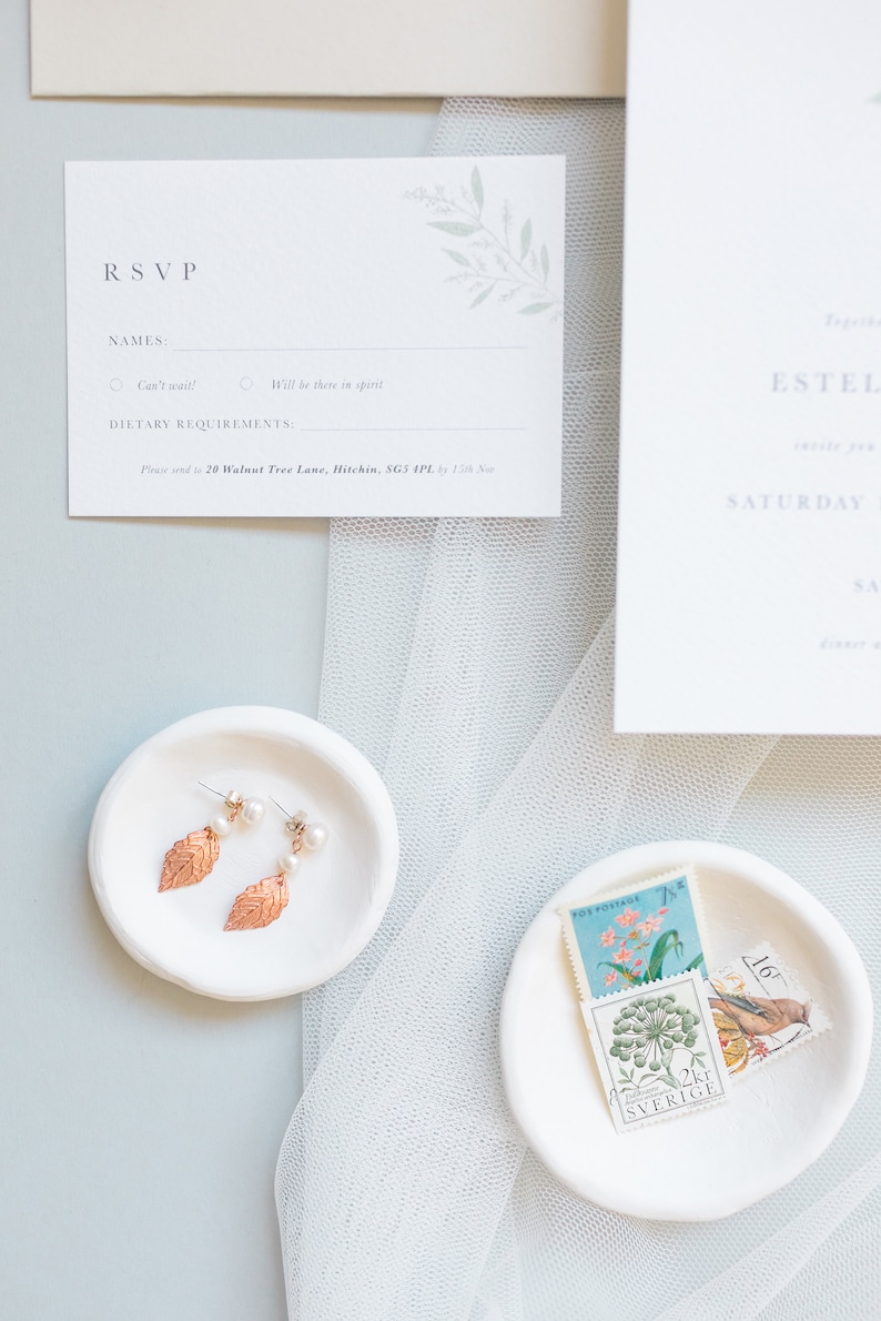 Wedding stationery flat lay with A7 rsvp card and part of 5x7 invitation. Design features minimal watercolour eucalyptus in pale green. Text is all in a classic serif font printed in grey on luxury white textured paper.