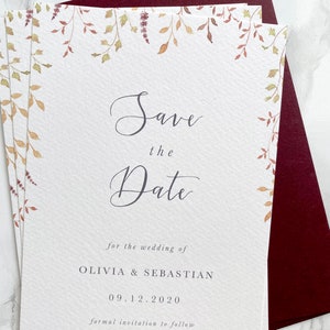 Pile of wedding save the dates with burgundy envelope. Autumn design with watercolour leaves coming down from top of the card to frame the text. Save the date is in modern calligraphy with serif text below. Printed on luxury textured card.