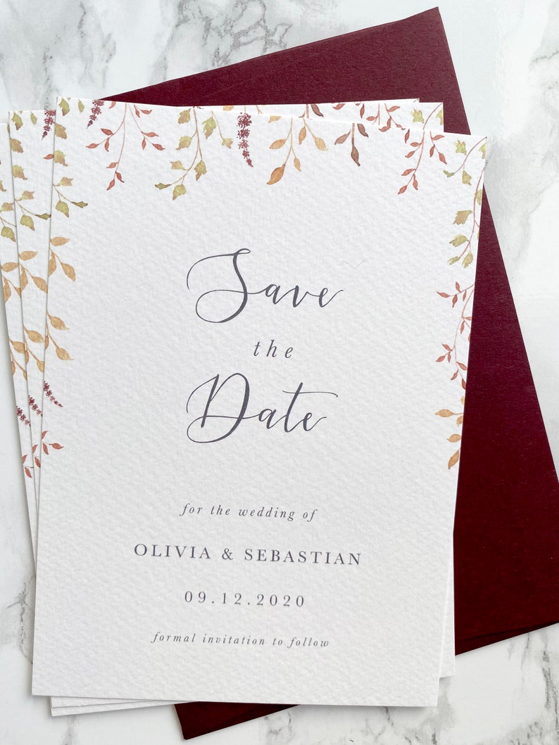 Pile of wedding save the dates with burgundy envelope. Autumn design with watercolour leaves coming down from top of the card to frame the text. Save the date is in modern calligraphy with serif text below. Printed on luxury textured card.