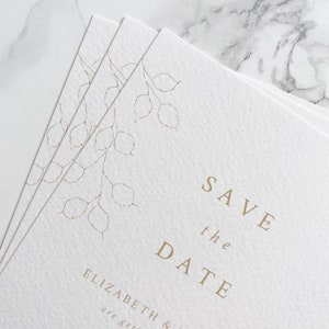 A small pile of wedding save the date cards with a close up of the botanical illustration in top left hand corner. Minimal and modern text is centred. Printed in pale gold on white luxury textured card.
