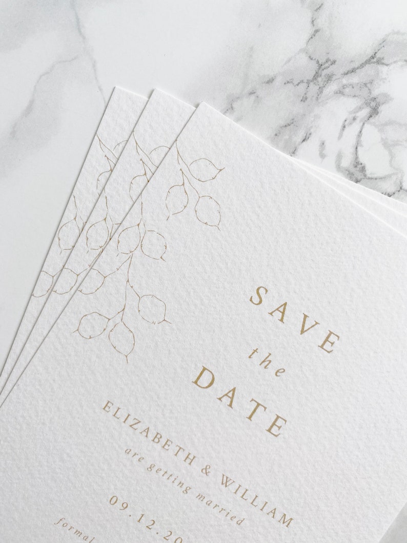 A small pile of wedding save the date cards with a close up of the botanical illustration in top left hand corner. Minimal and modern text is centred. Printed in pale gold on white luxury textured card.