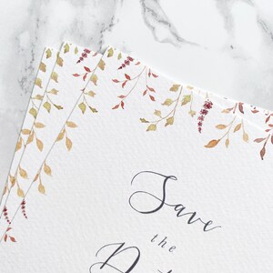 Close up of save the date pile with a focus on the watercolour illustrations. Illustrations are of autumn leaves in burnt orange, burgundy and gold. Save the date is written in modern calligraphy. Design is printed on luxury textured card stock