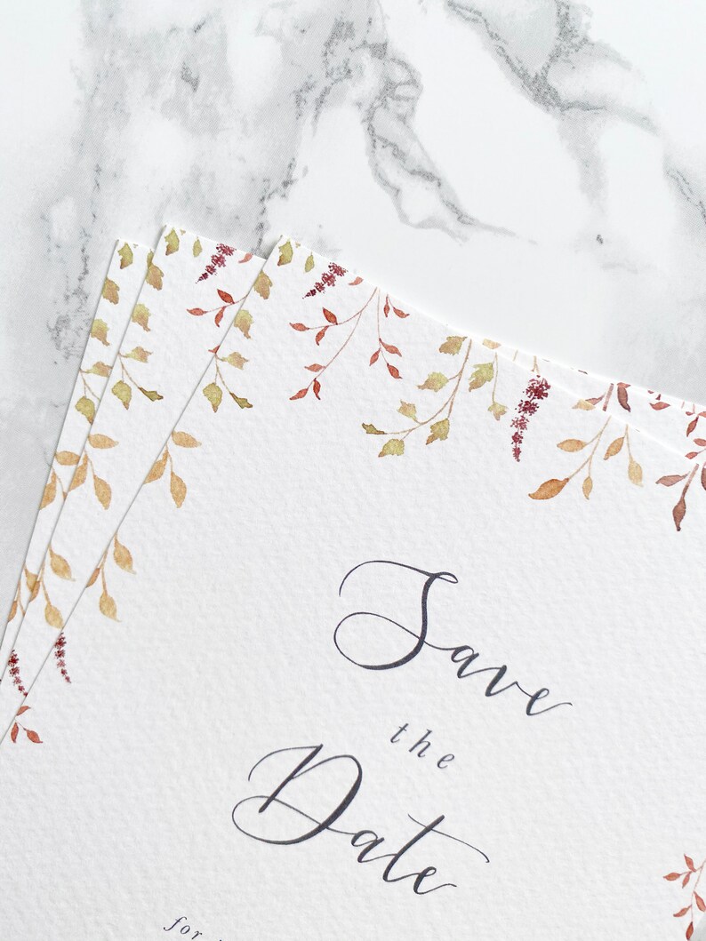 Close up of save the date pile with a focus on the watercolour illustrations. Illustrations are of autumn leaves in burnt orange, burgundy and gold. Save the date is written in modern calligraphy. Design is printed on luxury textured card stock