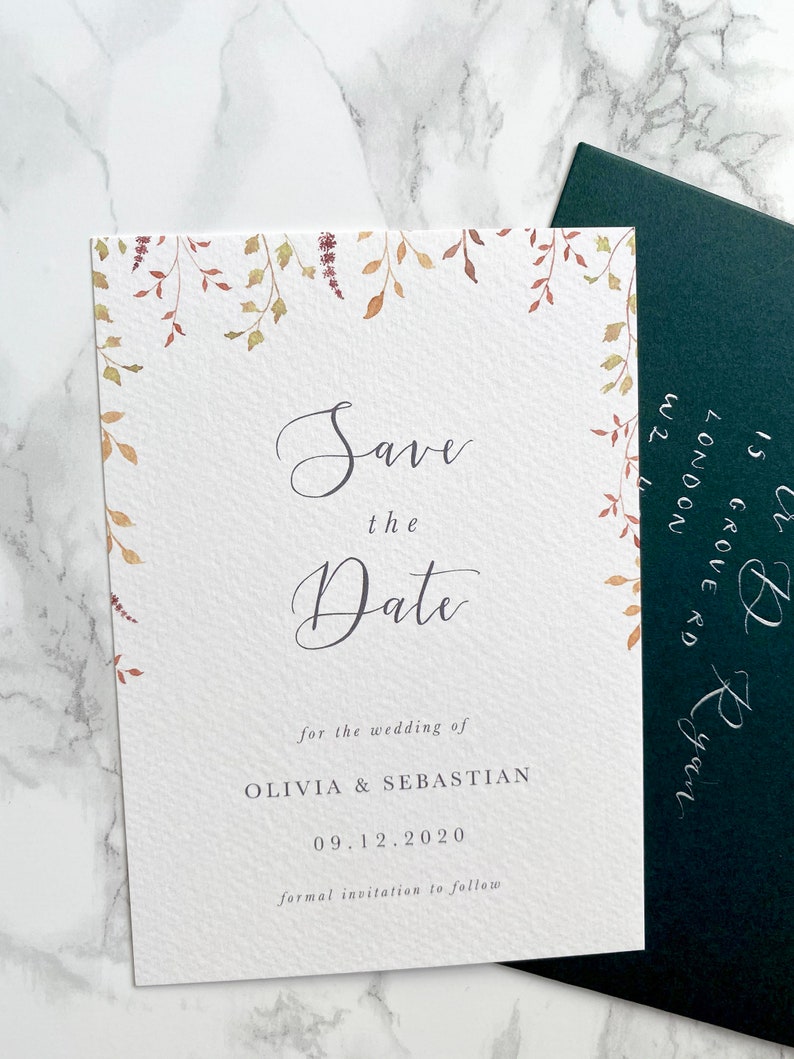 Wedding save the date with dark green envelope addressed in silver calligraphy. Autumn design with watercolour leaves coming down from top of the card to frame the text. Save the date is in modern calligraphy with serif text below.