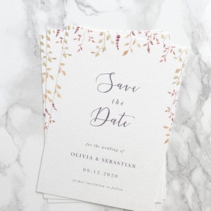 Full shot of a small pile of save the date cards for an autumn wedding. Design features watercolour autumn leaves at top with modern calligraphy script and elegant serif text below printed in grey on white, luxury card stock
