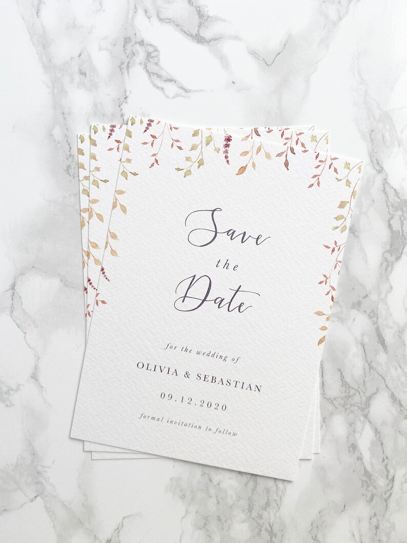 Full shot of a small pile of save the date cards for an autumn wedding. Design features watercolour autumn leaves at top with modern calligraphy script and elegant serif text below printed in grey on white, luxury card stock
