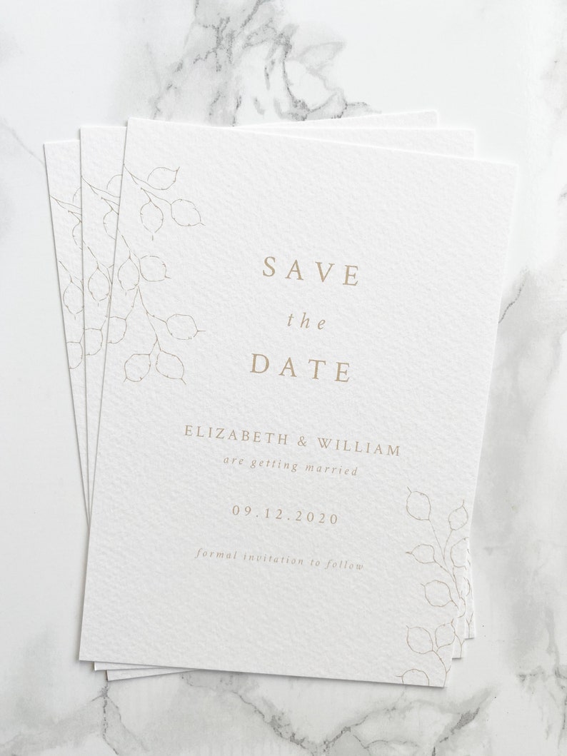 Pile of save the date cards for autumn or winter wedding. Minimal design with hand drawn lunaria branches and clean serif font design. Printed in pale gold onto white, luxury textured paper.