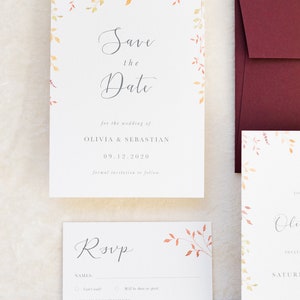 Wedding stationery flat lay with save the date and burgundy envelopes and gold wax seal. Design has watercolour leaves in autumn colours around the top edge and sides. The text is printed in grey on luxury white, textured paper.
