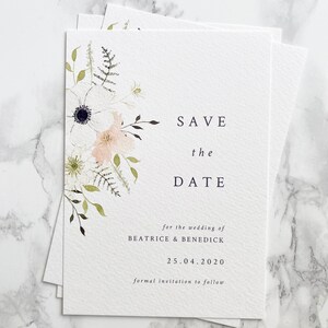 A6 portrait save the date cards with spring floral design painted in watercolour. Colours are peach, white and cream with various shades of green foliage. Text is minimal and modern in a classic serif font, aligned right.