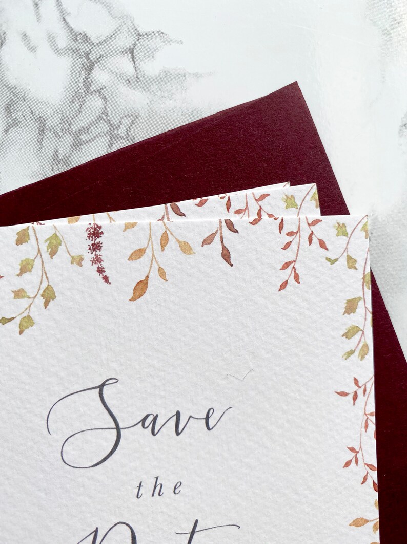 Close up of watercolour autumn leaves on save the date with burgundy envelope. Illustrations of different autumn foliage in burnt orange, burgundy and gold.