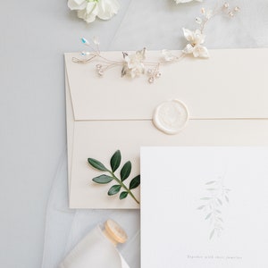 Wedding stationery flat lay with 5x7 inch invitation and neutral stone straight flap envelope with white wax seal. Design features pale green branch at top painted in watercolour with wedding details placed below in grey classic serif font.