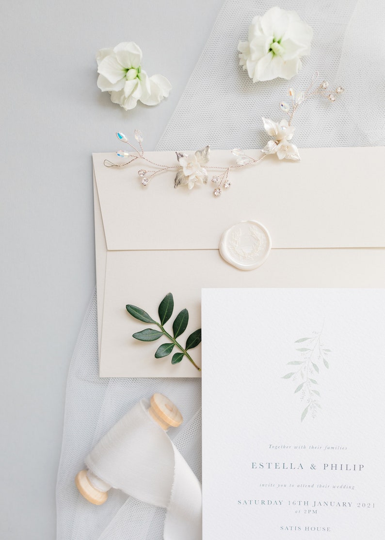 Wedding stationery flat lay with 5x7 inch invitation and neutral stone straight flap envelope with white wax seal. Design features pale green branch at top painted in watercolour with wedding details placed below in grey classic serif font.