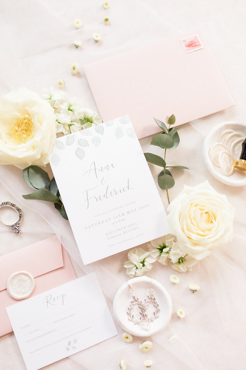 Wedding stationery suite flat lay with A6 invitation, rsvp card, blush envelopes and white wax seal. Design features watercolour eucalyptus in pale green with calligraphy names in grey. Printed on fine art textured white paper.