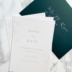 Pile of save the date cards for autumn or winter wedding with dark green envelope and silver calligraphy addressing. Minimal design with hand drawn lunaria branches and clean serif font design. Printed in pale gold onto white, luxury textured paper.