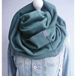 Könnte beinhalten: Ein teal-grüner Infinity-Schal aus weichem Stoff, der auf einer Schaufensterpuppe präsentiert wird. Der Schal hat eine Tasche mit einem kleinen, rechteckigen Aufnäher. Der Schal ist über einer hellblauen Jeansjacke drapiert.