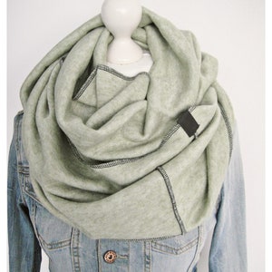 Könnte beinhalten: Hellgrüner Loop-Schal mit schwarzen Nähten und einem kleinen schwarzen Etikett. Der Schal ist über einer Jeansjacke drapiert. Der Schal wirkt weich und gemütlich, ideal für kühlere Tage. Der Schal ist etwa 150 cm lang.