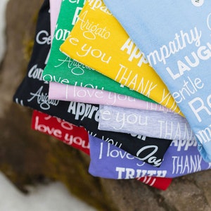 May include: A stack of t-shirts with different colors and positive messages printed on them. The messages include "Arigato", "Thank You", "Love You", "Appreciation", "Joy", "Truth", "Empathy", "Laugh", "Gentle", "Respect", and "Compassion".