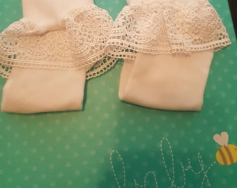 May include: A pair of white baby socks with delicate lace trim. The socks are displayed on a teal background with the word "baby" written in cursive. A yellow and white bee graphic is also present.