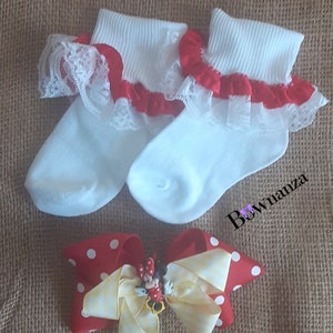 May include: A pair of white baby socks with red ruffled trim and lace detailing, paired with a red and white polka dot bow with a yellow striped ribbon and a Minnie Mouse embellishment. The text "Bownanza" is visible.