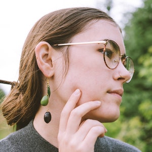 May include: A pair of dangle earrings with green and black oval-shaped beads suspended from gold-colored hooks. The earrings are worn by a person wearing a gray sweater and round eyeglasses with a tortoiseshell pattern.