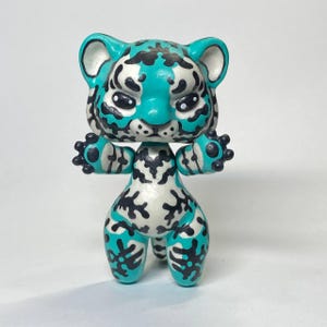 Op de afbeelding: Een klein, gestileerd tijgerfiguurtje met een gladde, glanzende afwerking. Het wezen heeft een levendige turquoise en witte basiskleur, geaccentueerd met opvallende, handgeschilderde zwarte strepen en vlekken op de kop, romp en ledematen.