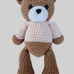 Könnte beinhalten: Ein handgehäkelter brauner Teddybär mit weißer Schnauze und schwarzen Augen. Der Bär trägt einen hellrosa Pullover. Die Arme und Beine des Bären hängen herunter, vor einem hellgrauen Hintergrund.