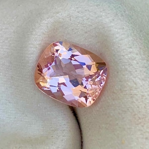 May include: A faceted, peach-coloured gemstone with a complex internal structure. The gem is set against a white cloth, with the text "RESEARCH SWISS LAB" visible in the lower right corner. The stone's shape is irregular, with multiple facets catching the light.